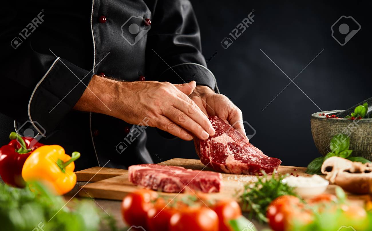 Chef seasoning a thick raw beef steak with spice rub on a wooden board with fresh vegetables and herbs in a close up of the hands - 115257912 Chef seasoning a thick raw beef steak with spice rub on a wooden board with fresh vegetables and herbs in a close up of the hands - 115257912