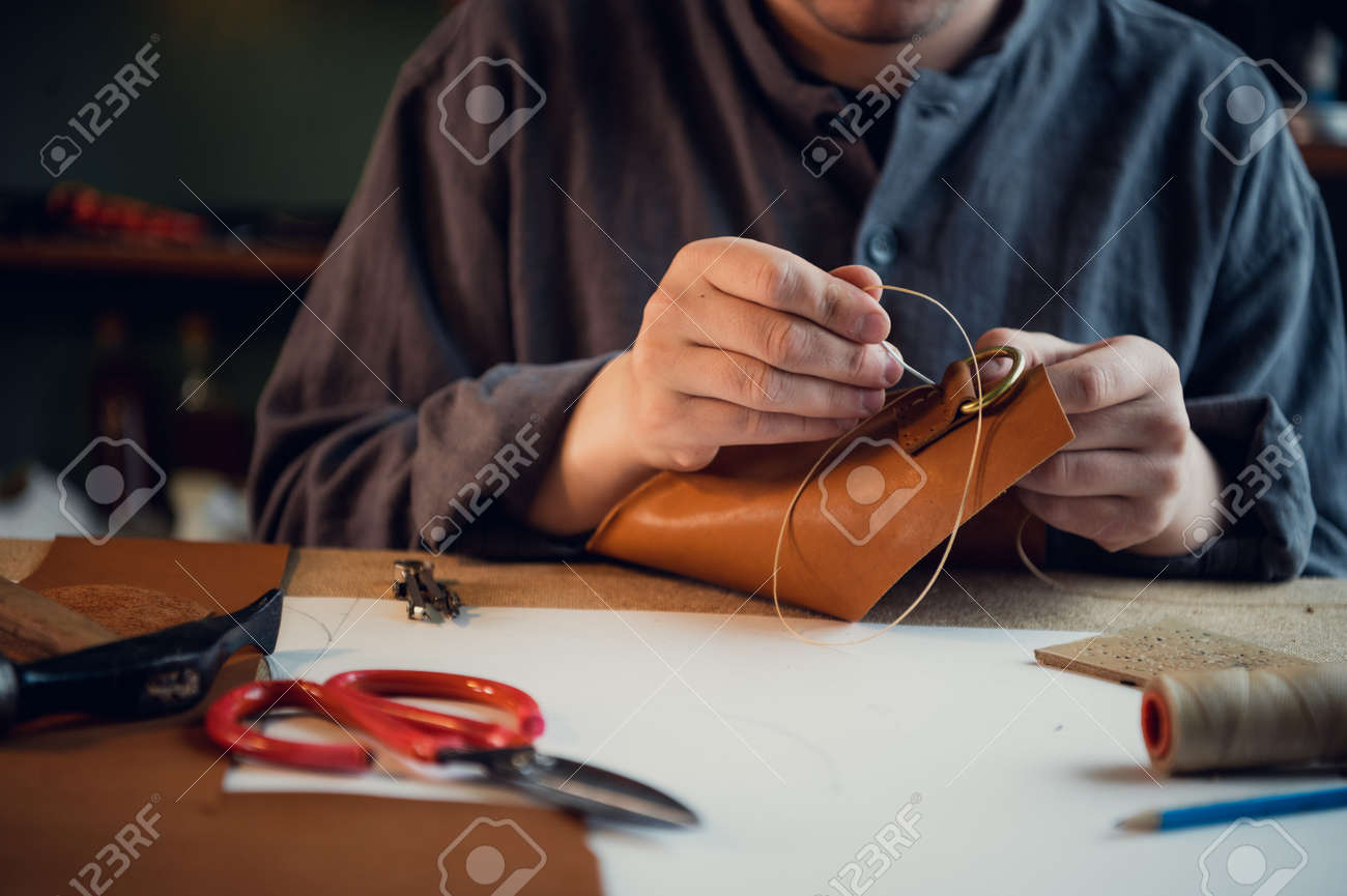 Sitting at a table in the workshop a young guy manually sews leather elements to each other. - 164872236 Sitting at a table in the workshop a young guy manually sews leather elements to each other. - 164872236