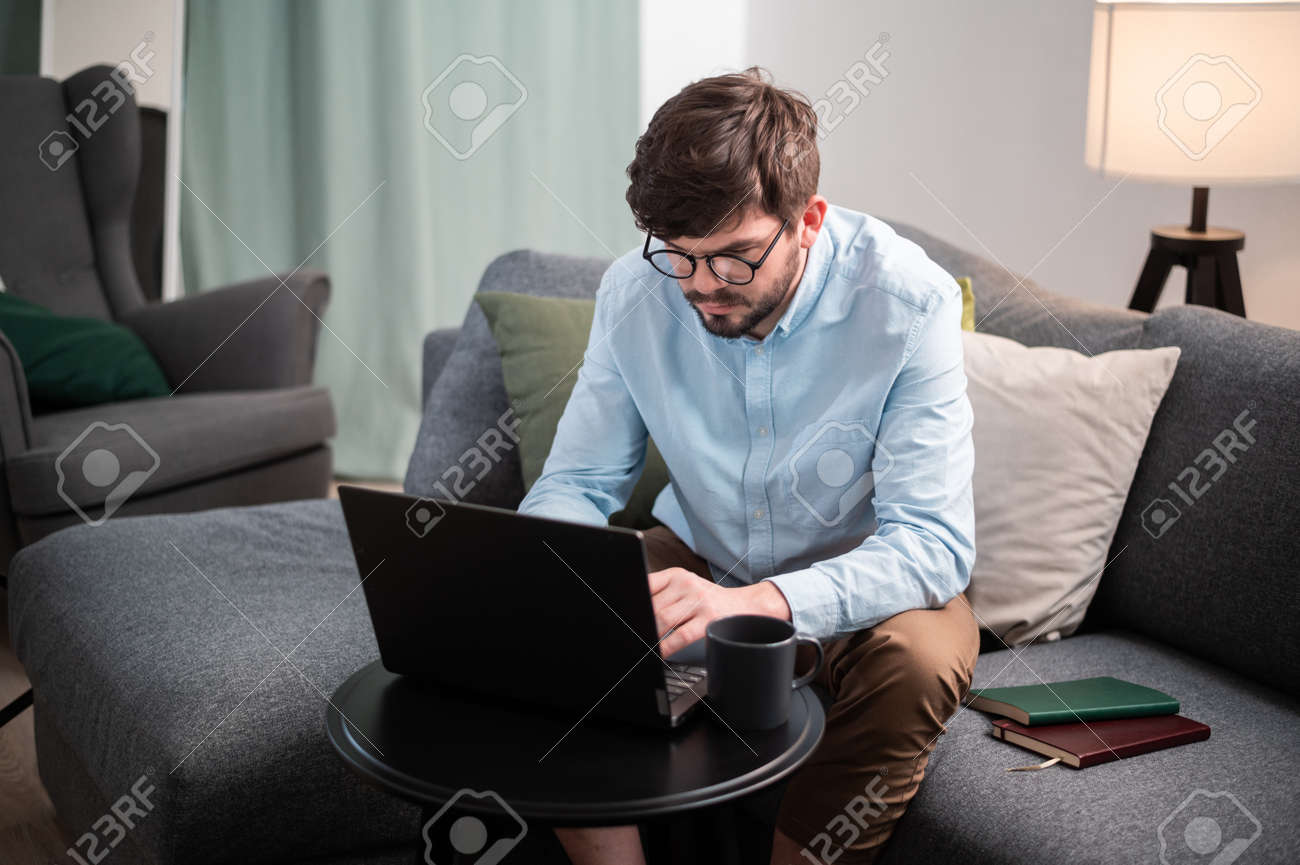 Hard work with the help of a young mans laptop, he sits on the couch, makes a report and drinks an invigorating coffee - 170401089 Hard work with the help of a young mans laptop, he sits on the couch, makes a report and drinks an invigorating coffee - 170401089