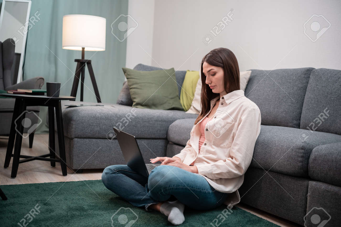 A young girl working from home, she sits on the floor with a laptop on the mat next to the bed during the covid-19 quarantine - 170401094 A young girl working from home, she sits on the floor with a laptop on the mat next to the bed during the covid-19 quarantine - 170401094