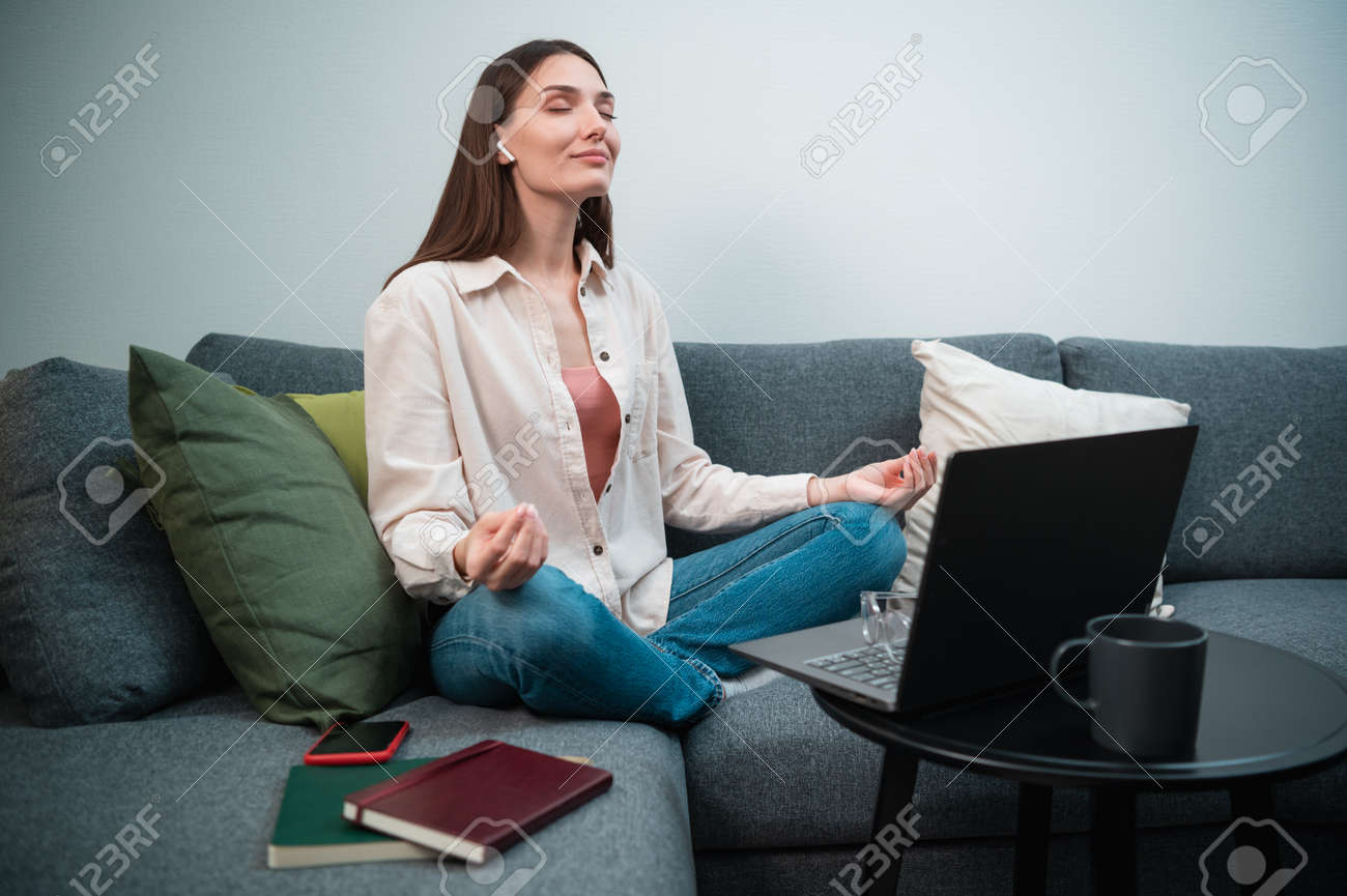 At lunchtime, the manager woman relieves stress with the help of the lotus asana, sitting at home on the couch - 170401097 At lunchtime, the manager woman relieves stress with the help of the lotus asana, sitting at home on the couch - 170401097