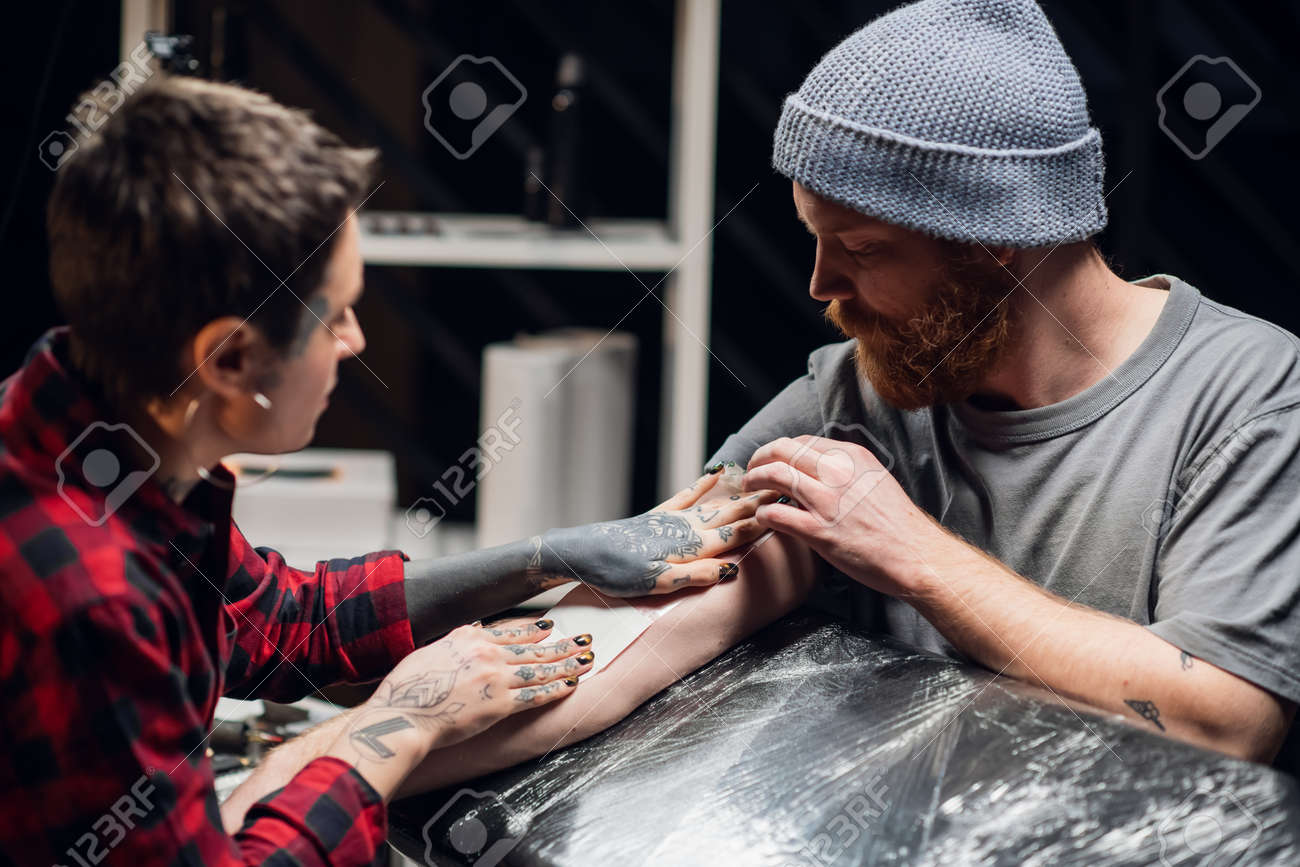 Girl tattoo artist takes care of the young mans hand after tattooing. Makes the dressing transparent film with the application of a moisturizer. - 170401059 Girl tattoo artist takes care of the young mans hand after tattooing. Makes the dressing transparent film with the application of a moisturizer. - 170401059