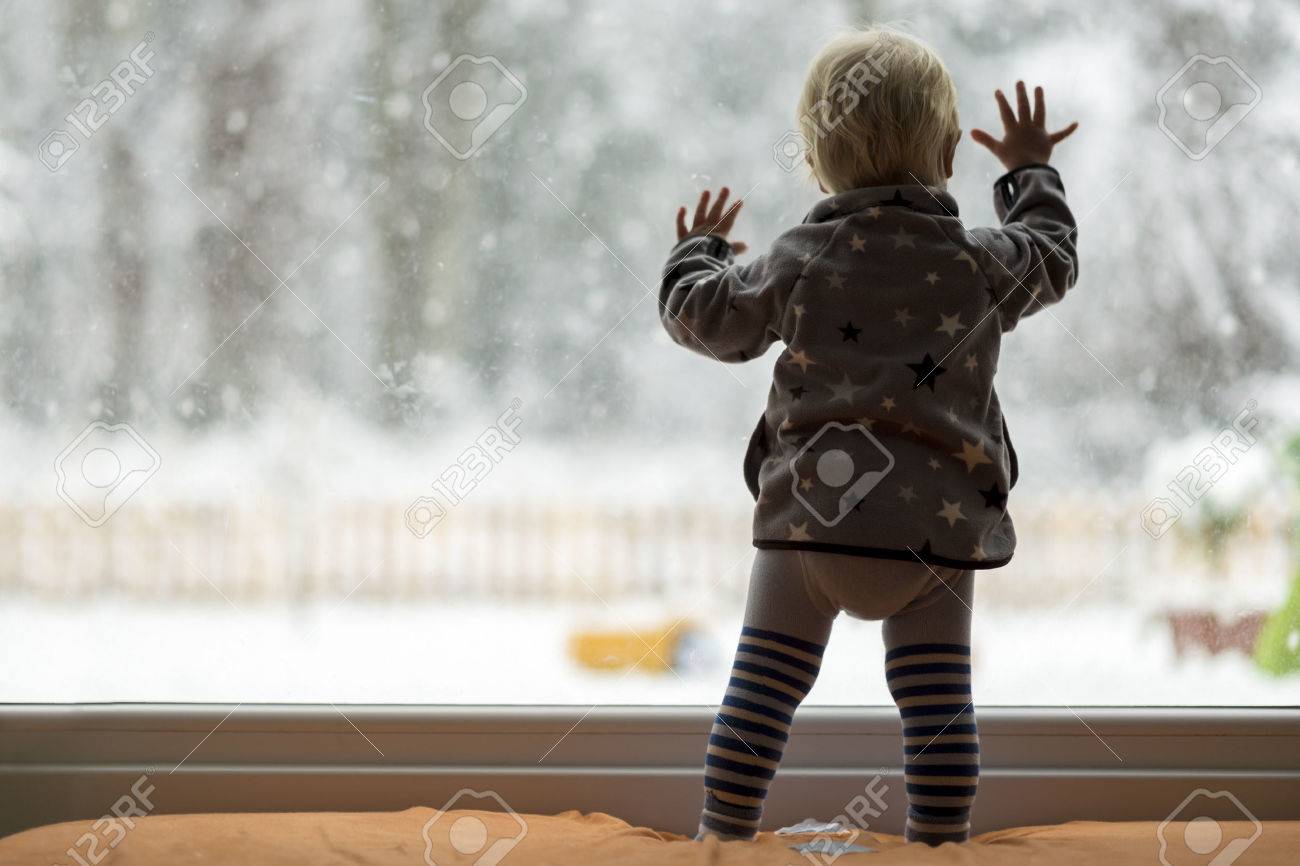 View form behind of toddler child standing in front of a big window leaning against it looking outside at a snowy nature. - 50415994 View form behind of toddler child standing in front of a big window leaning against it looking outside at a snowy nature. - 50415994