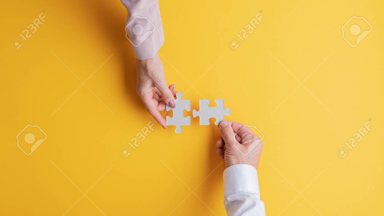 Top view of male and female hands joining two matching puzzle pieces together in a conceptual image. Over yellow background. - 131604808 Top view of male and female hands joining two matching puzzle pieces together in a conceptual image. Over yellow background. - 131604808