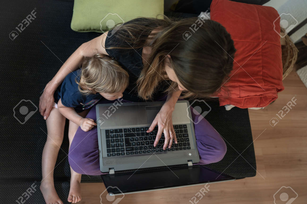 Top view of a young mother sitting on a sofa with laptop computer on her knees working with her toddler daughter cuddling next to her. - 169239360 Top view of a young mother sitting on a sofa with laptop computer on her knees working with her toddler daughter cuddling next to her. - 169239360