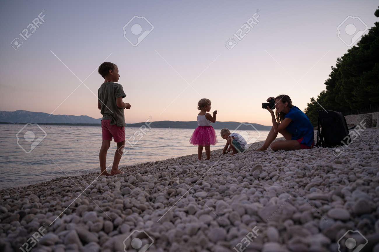 Young mother photographing her three children playing on pebble beach in a beautiful summer evening. - 169239412 Young mother photographing her three children playing on pebble beach in a beautiful summer evening. - 169239412