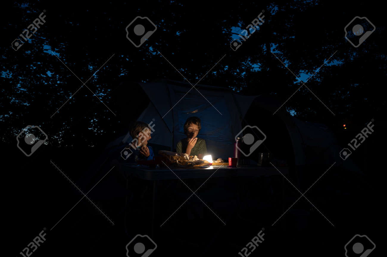 Two brothers sitting by their tent in a camping place eating dinner by a dimmed light. - 171940301 Two brothers sitting by their tent in a camping place eating dinner by a dimmed light. - 171940301