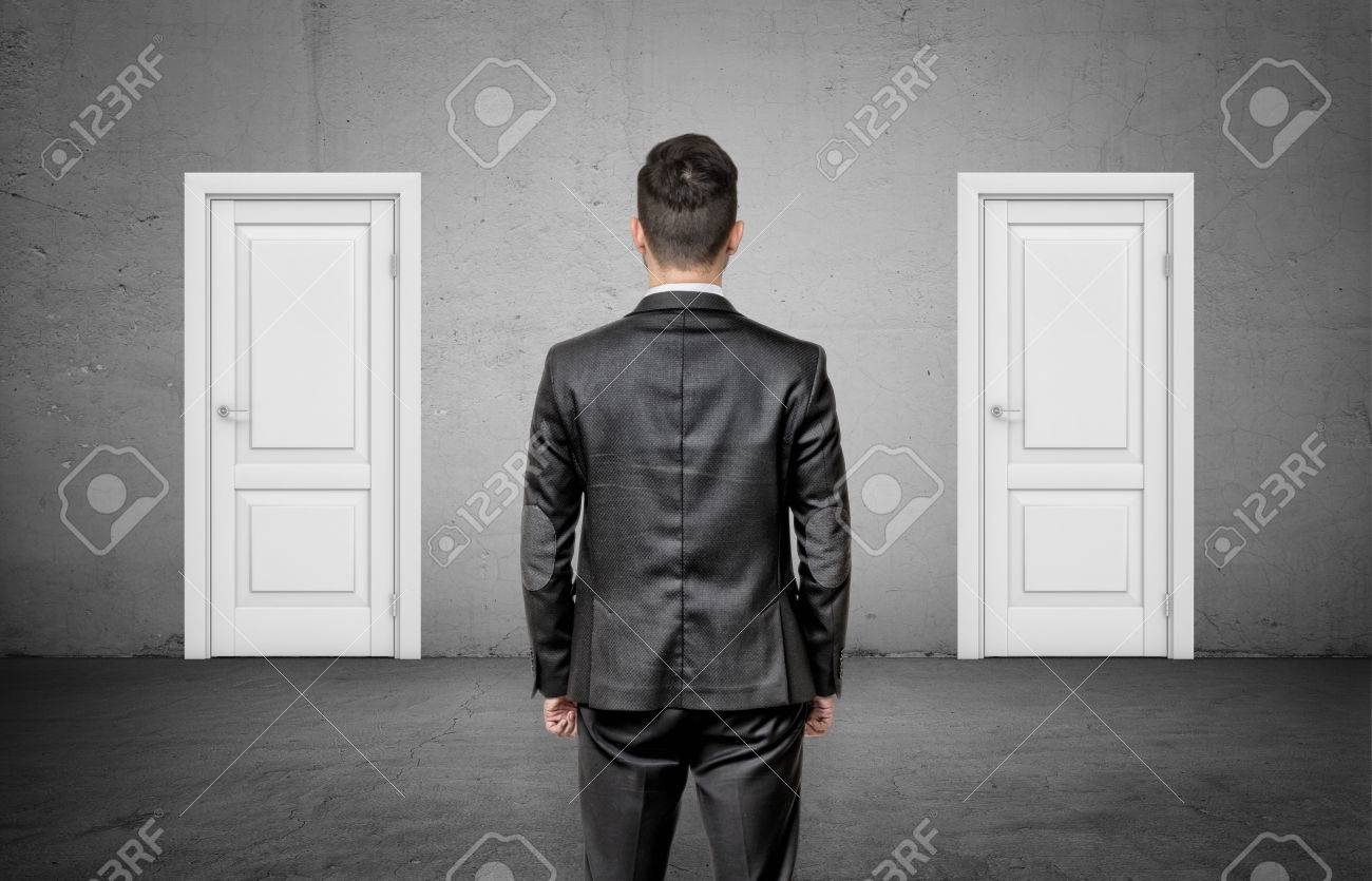 A businessman with his back turned stands between two identical closed white doors. - 85241147 A businessman with his back turned stands between two identical closed white doors. - 85241147