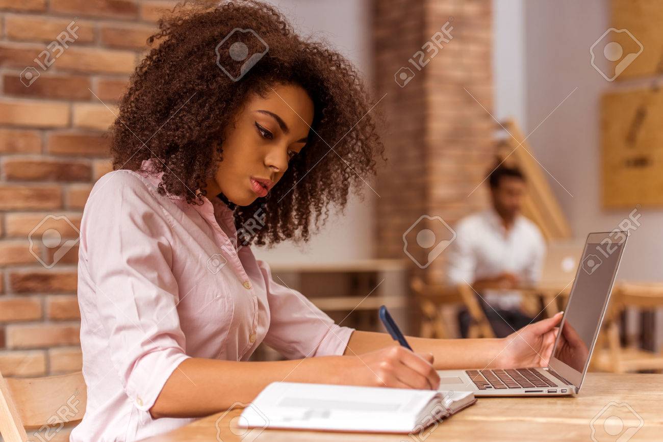 Young beautiful Afro-American businesswoman using laptop and writing in notebook while studying in cafe - 51766485 Young beautiful Afro-American businesswoman using laptop and writing in notebook while studying in cafe - 51766485