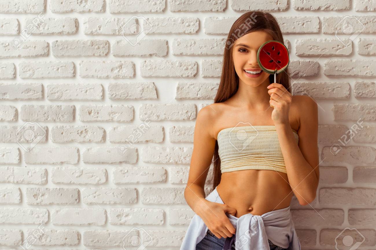 Linda Adolescente En Un Top Y Pantalones Cortos Cubre Un Ojo Con Una Piruleta, Mirando En La Cámara Y Sonriendo, De Pie Contra La Pared De Ladrillo Blanco Fotos, Retratos, Imágenes