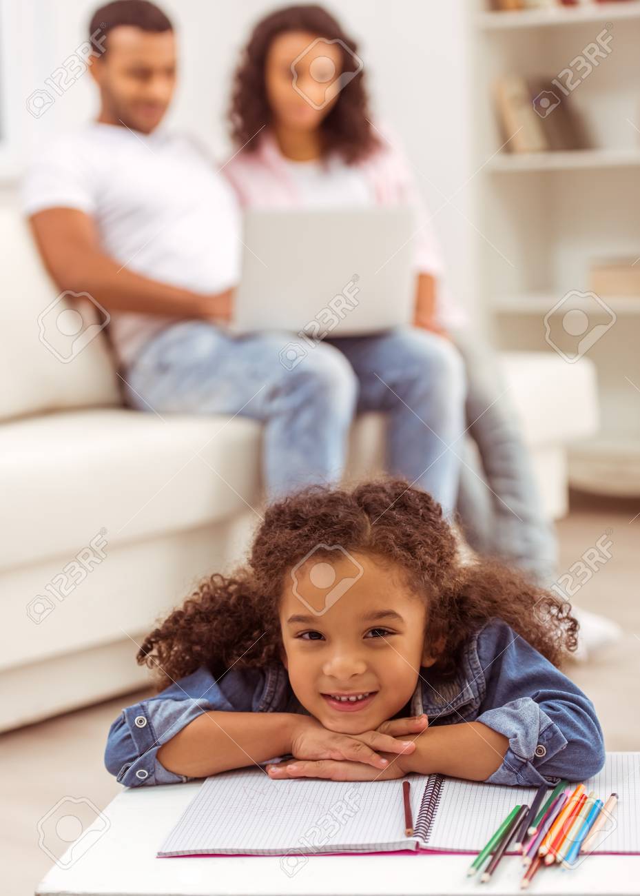 かわいい少しのアフリカ系アメリカ人の女の子は彼女の両親は ラップトップを使用してバック グラウンドで 部屋の床の上に横たわる の写真素材 画像素材 Image