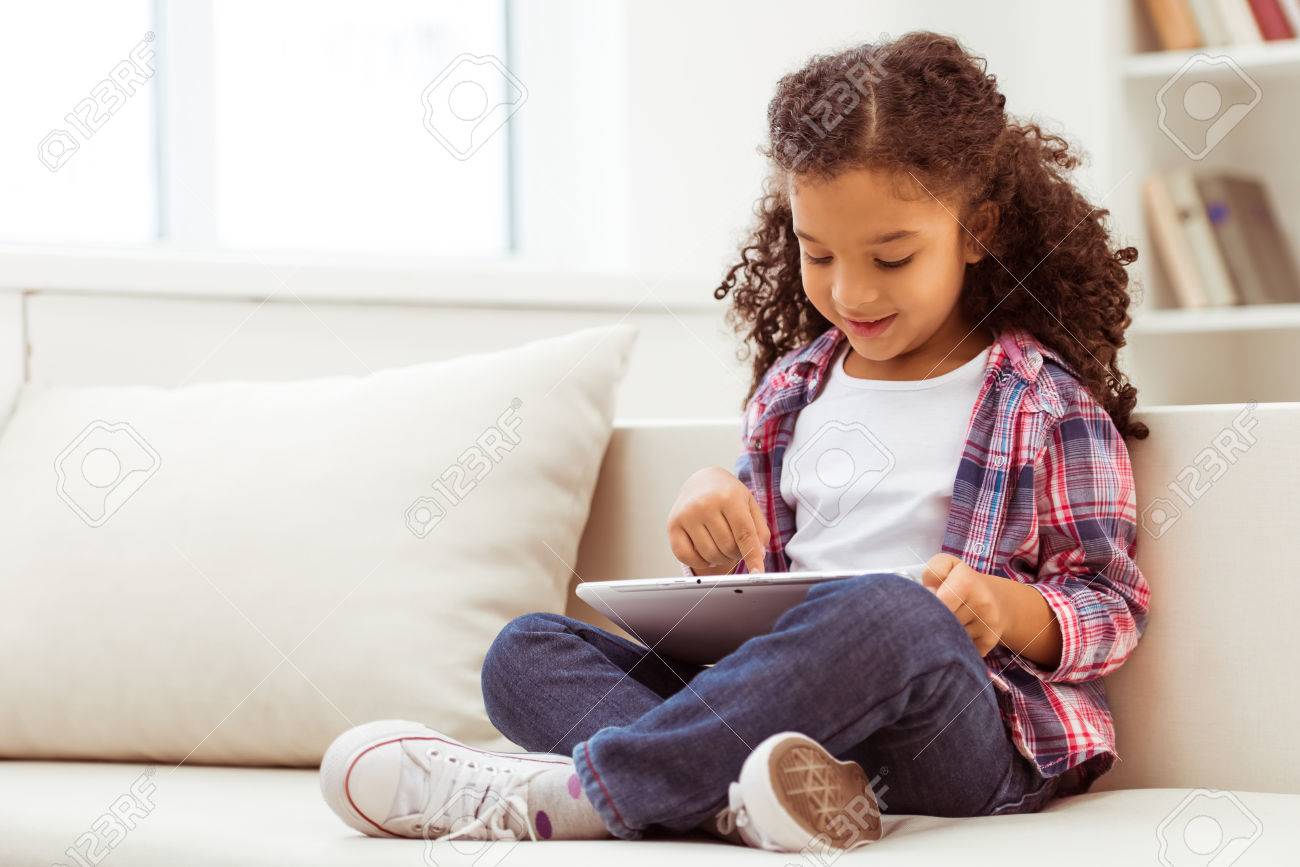 タブレットを使用して 部屋のソファに腰掛けながら笑顔のカジュアルな服でかわいい小さなアフリカ系アメリカ人女の子 の写真素材 画像素材 Image