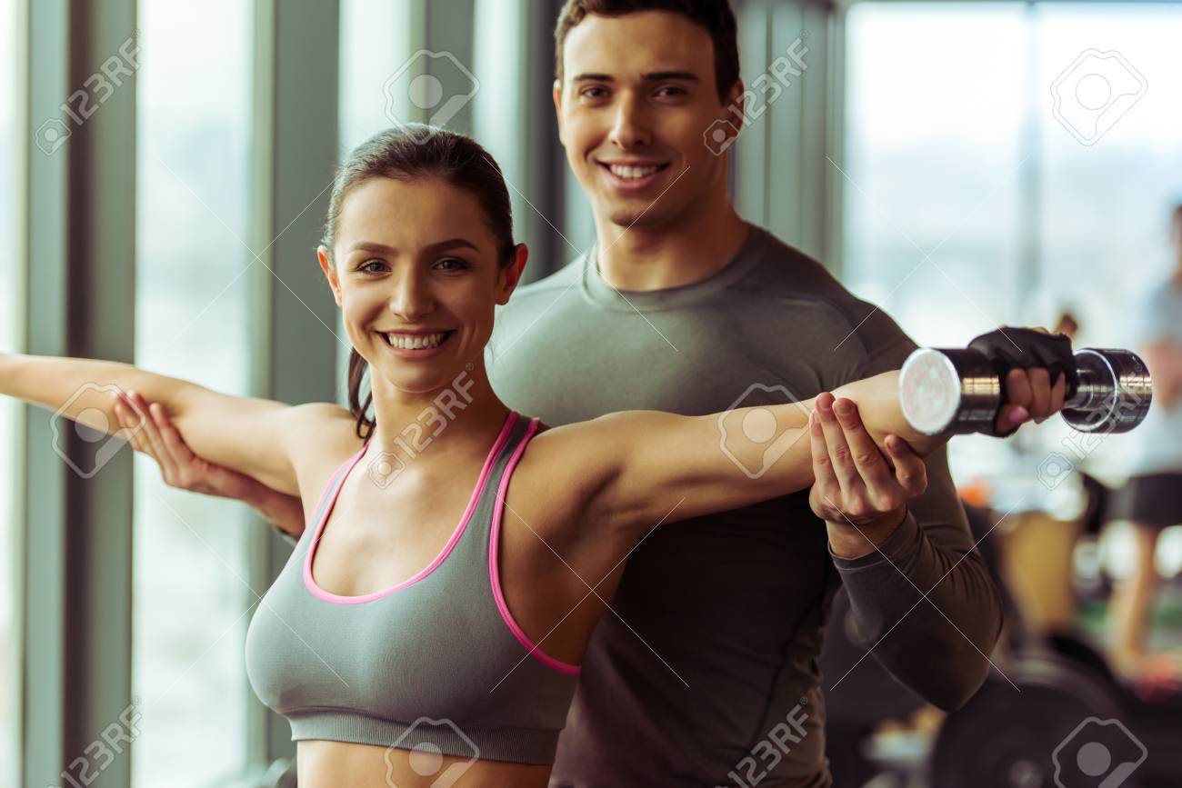 彼女を助けるハンサムな筋肉トレーナーのジムでダンベル扱う魅力的な若い女性 の写真素材 画像素材 Image