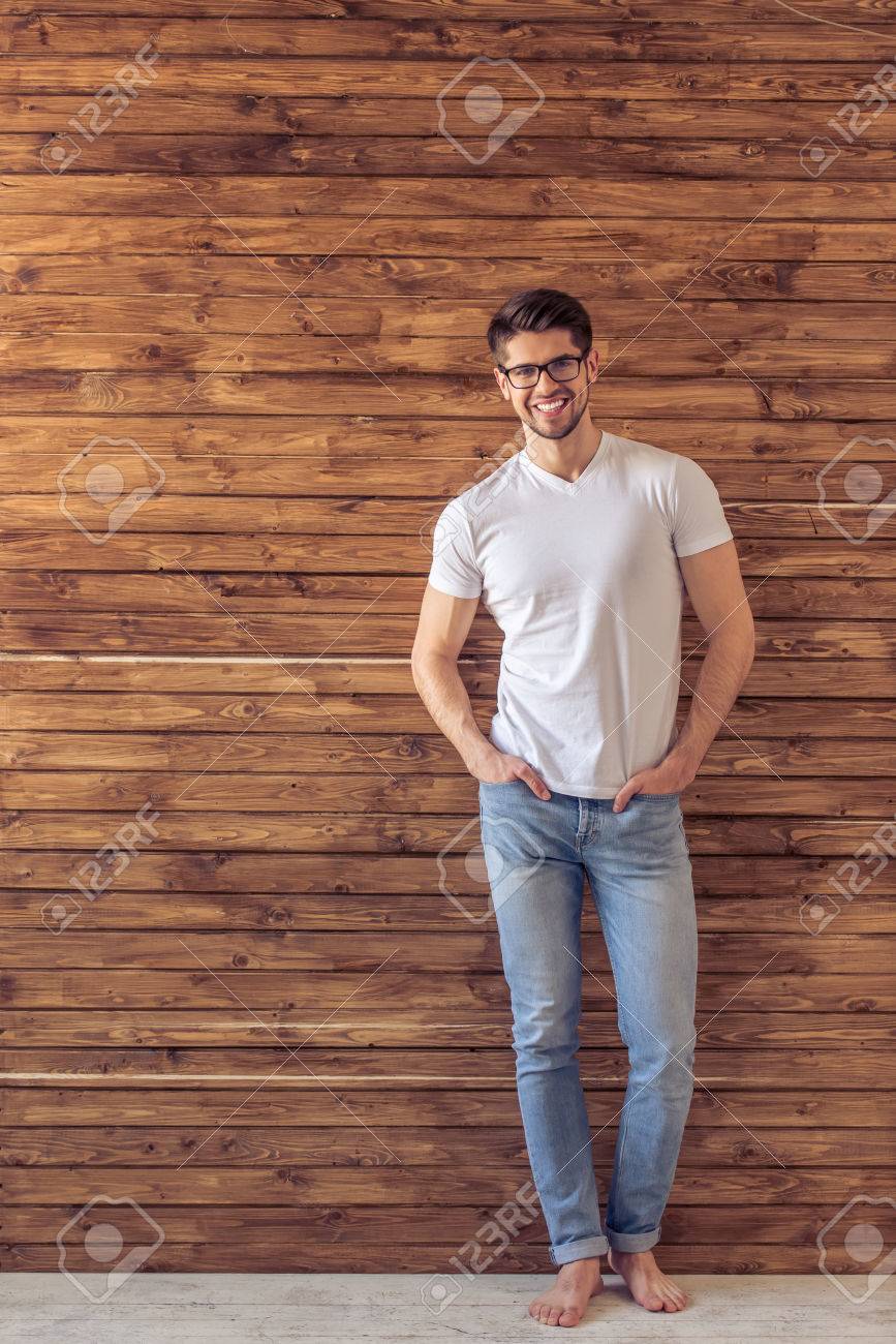 De Cuerpo Entero De Un Hombre Guapo En Ropa Casual Y Gafas Mirando Cámara Y Sonriendo, De Pie Manos En Los Bolsillos Contra La Pared De Madera Fotos,