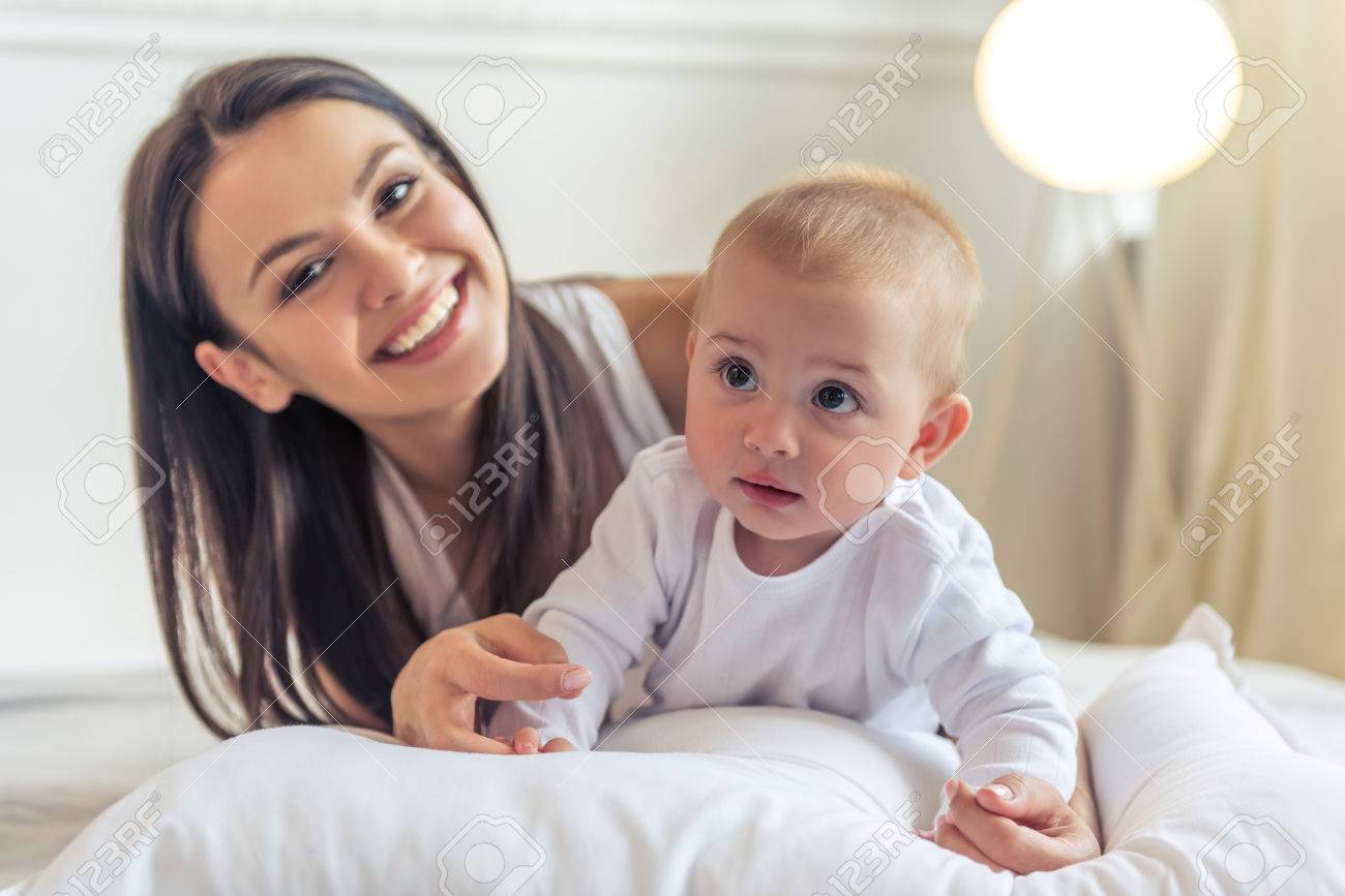 美しい若いお母さんの肖像画 彼女の甘い小さな赤ちゃんのカメラを見て 笑って 自宅のベッドに横になっています の写真素材 画像素材 Image
