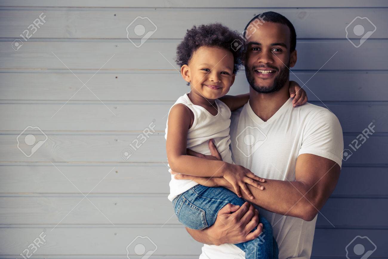 Portrait of handsome young Afro American father and his cute child hugging, looking at camera and smiling - 61557227 Portrait of handsome young Afro American father and his cute child hugging, looking at camera and smiling - 61557227