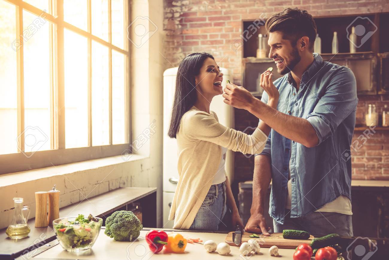 Beautiful young couple is feeding each other and smiling while cooking in kitchen at home - 64346720 Beautiful young couple is feeding each other and smiling while cooking in kitchen at home - 64346720