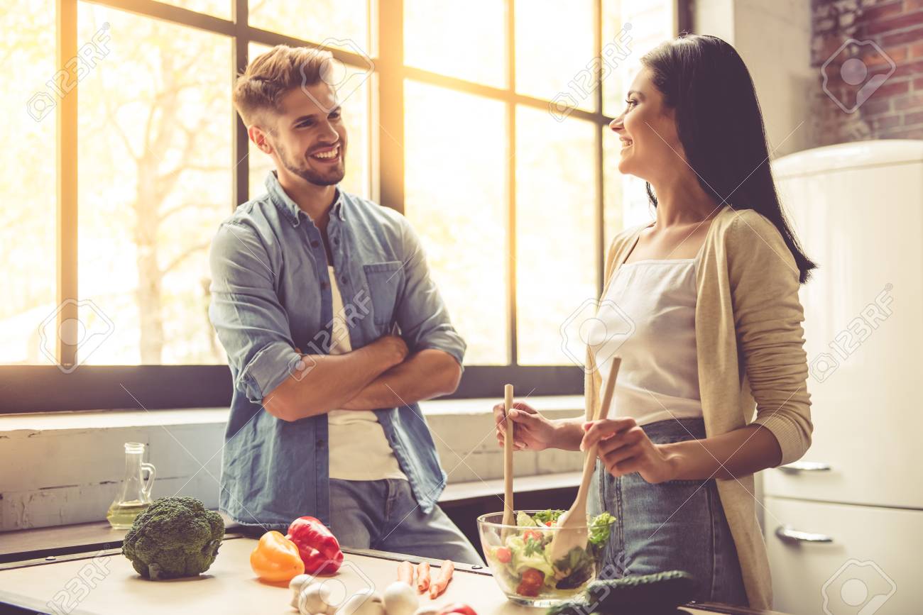 Beautiful young couple is talking, looking at camera and smiling while cooking in kitchen at home. Woman is mixing salad - 64346737 Beautiful young couple is talking, looking at camera and smiling while cooking in kitchen at home. Woman is mixing salad - 64346737