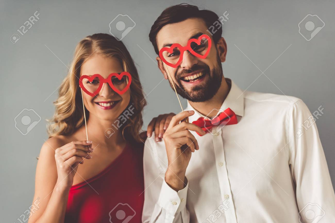 Beautiful elegant girl in red dress and guy in classic shirt and red bow tie are holding a paper heart glasses and smiling, on gray background - 66793477 Beautiful elegant girl in red dress and guy in classic shirt and red bow tie are holding a paper heart glasses and smiling, on gray background - 66793477