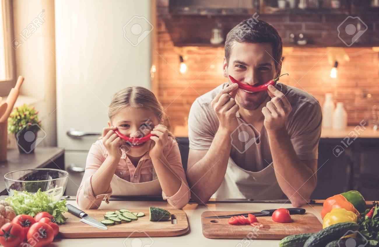 Cute little girl and her handsome dad are holding pepper and smiling while cooking in kitchen at home - 69038046 Cute little girl and her handsome dad are holding pepper and smiling while cooking in kitchen at home - 69038046