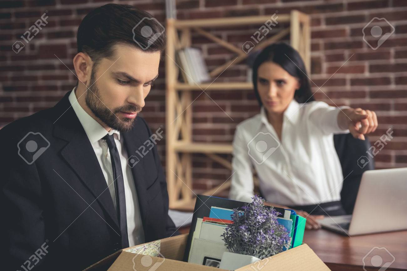 Getting fired. Handsome businessman in suit is holding a box with his stuff, woman in the background is pointing away - 72680907 Getting fired. Handsome businessman in suit is holding a box with his stuff, woman in the background is pointing away - 72680907