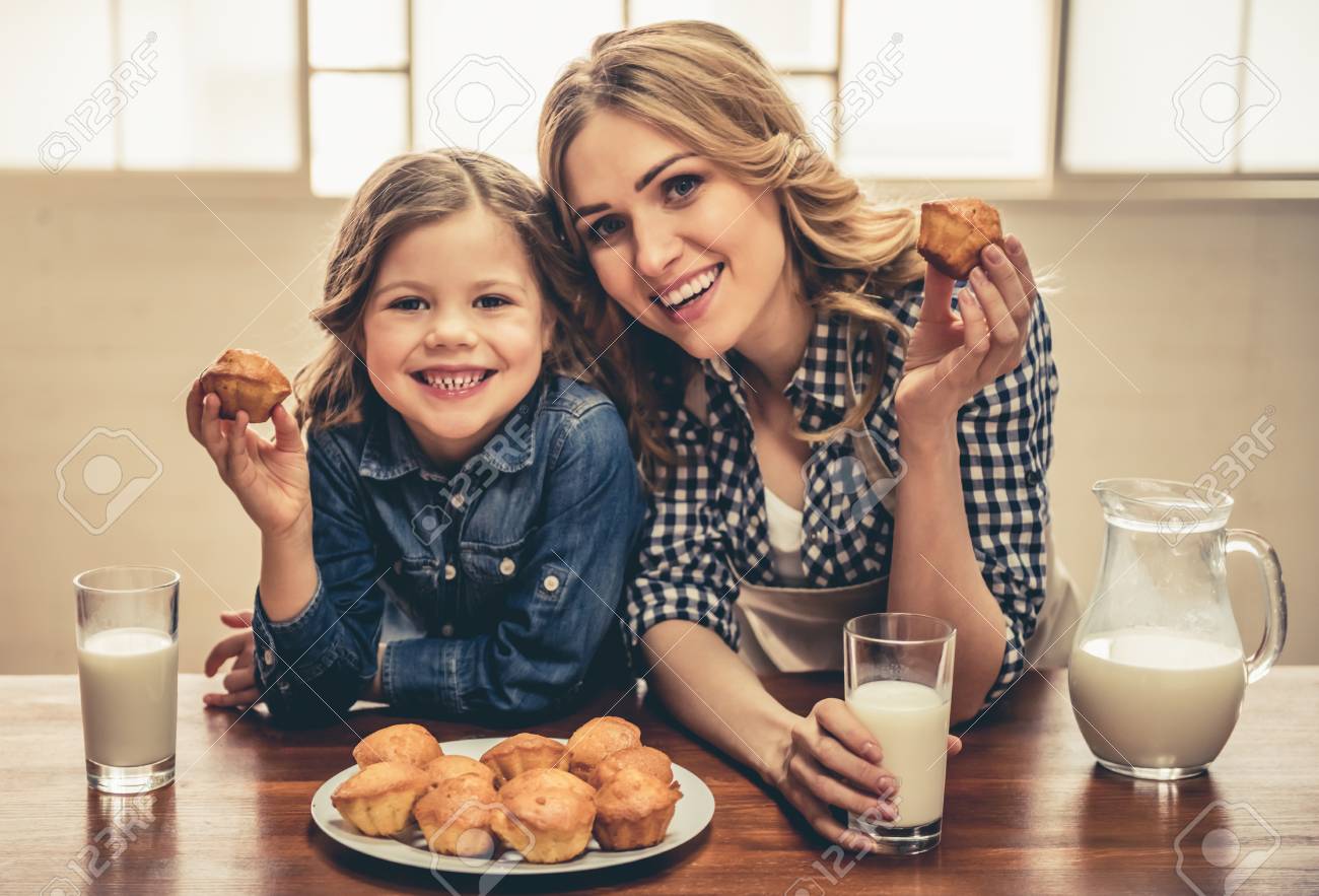Cute little girl and her beautiful young mom are eating muffins with milk, looking at camera and smiling while resting at home - 74330070 Cute little girl and her beautiful young mom are eating muffins with milk, looking at camera and smiling while resting at home - 74330070
