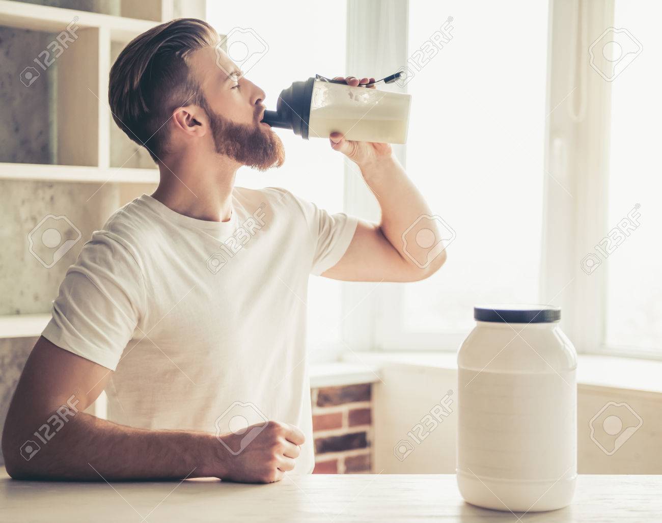 Handsome young bearded sportsman is drinking while preparing sport nutrition in kitchen at home - 79149126 Handsome young bearded sportsman is drinking while preparing sport nutrition in kitchen at home - 79149126