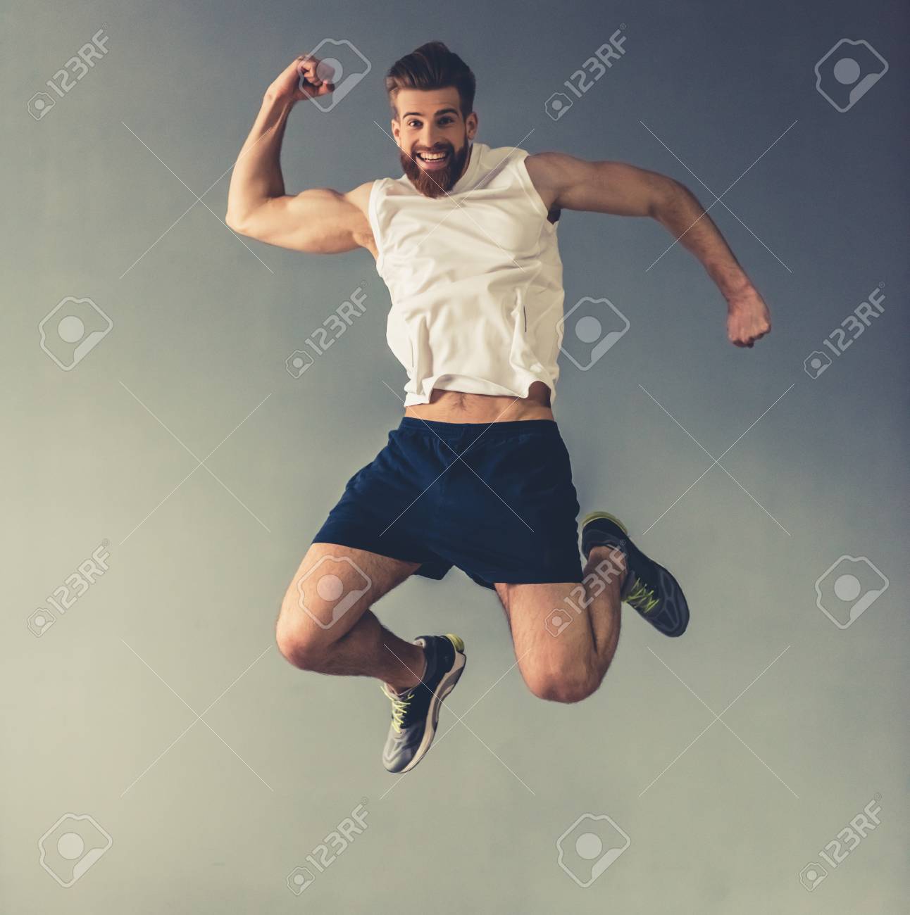 Handsome young bearded sportsman is showing muscles, jumping and smiling, on gray background - 79241600 Handsome young bearded sportsman is showing muscles, jumping and smiling, on gray background - 79241600