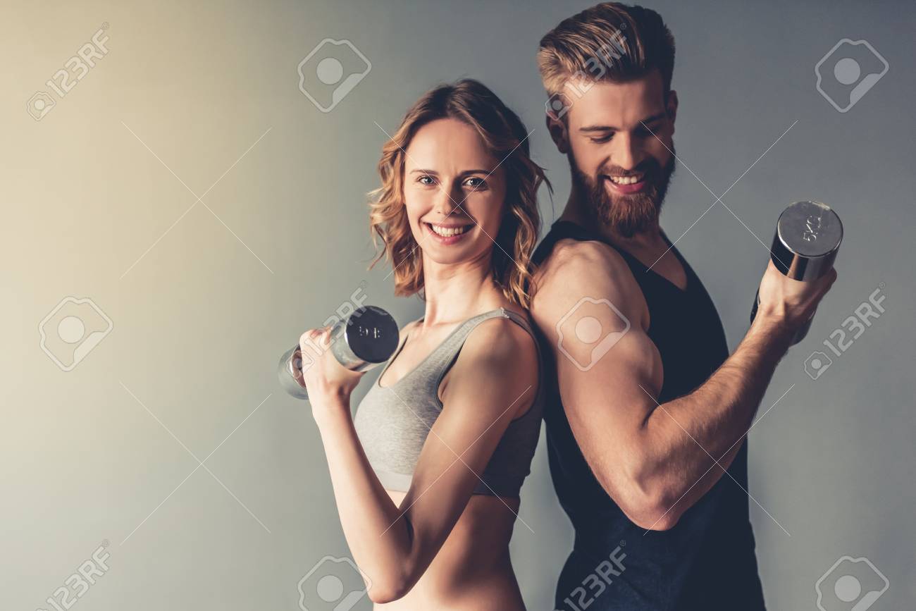 Beautiful young sports people are working out with dumbbells and smiling, on gray background - 79149180 Beautiful young sports people are working out with dumbbells and smiling, on gray background - 79149180