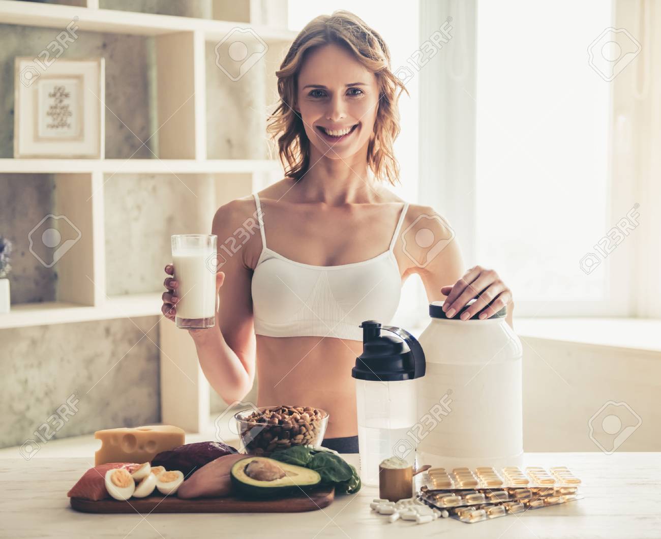 Beautiful young sportswoman is looking at camera and smiling while preparing sport nutrition in kitchen at home - 79149197 Beautiful young sportswoman is looking at camera and smiling while preparing sport nutrition in kitchen at home - 79149197