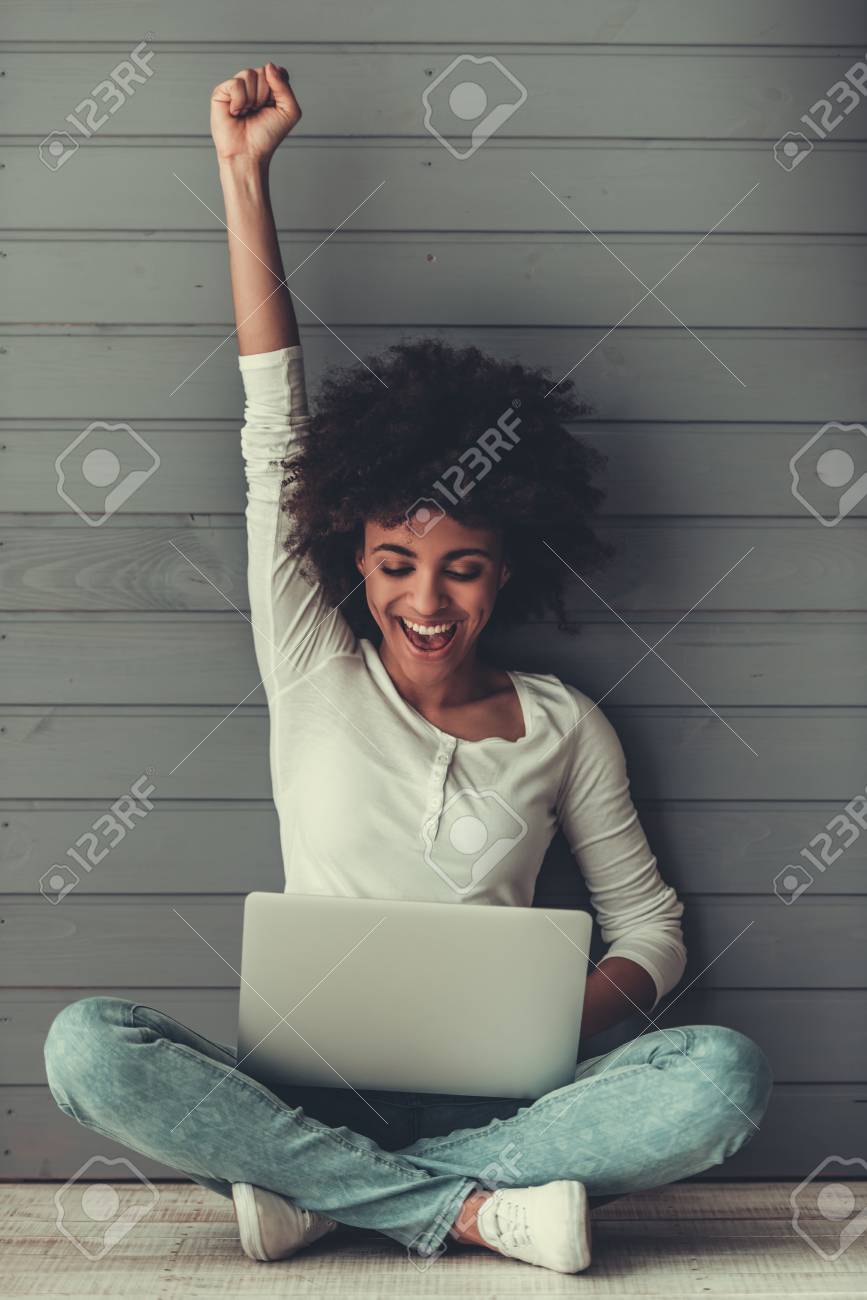 Attractive Afro American girl is using a laptop, raising hand in fist and smiling while sitting on the floor - 81369657 Attractive Afro American girl is using a laptop, raising hand in fist and smiling while sitting on the floor - 81369657