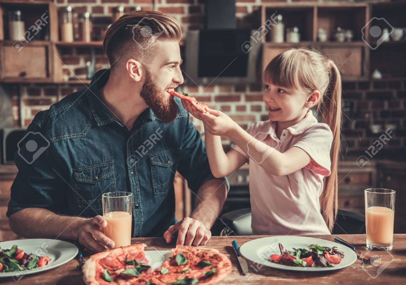 Cute little girl and her handsome bearded dad are smiling while drinking juice and eating pizza in kitchen - 82314666 Cute little girl and her handsome bearded dad are smiling while drinking juice and eating pizza in kitchen - 82314666