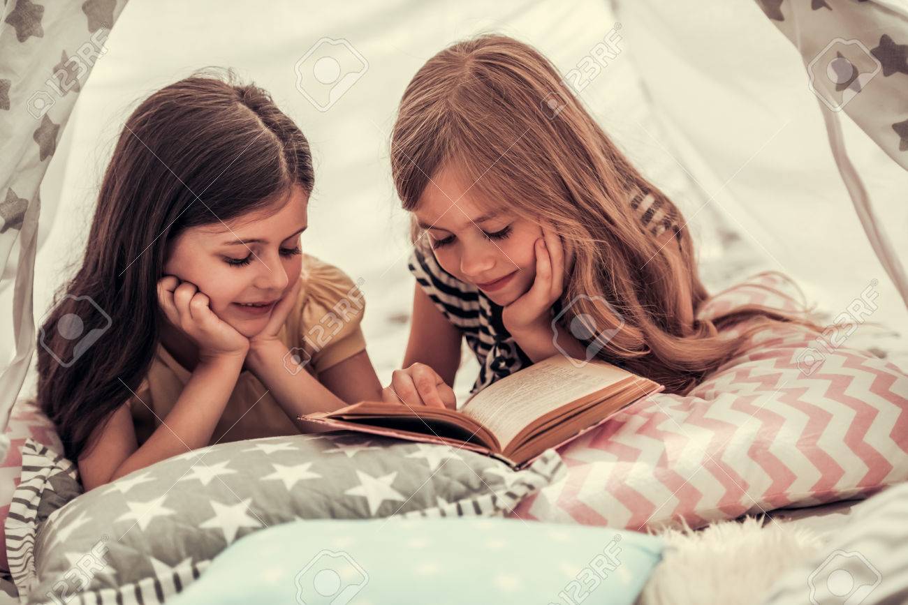 Two cute little girls are reading a book and smiling while playing together in child's teepee - 84790756 Two cute little girls are reading a book and smiling while playing together in child's teepee - 84790756