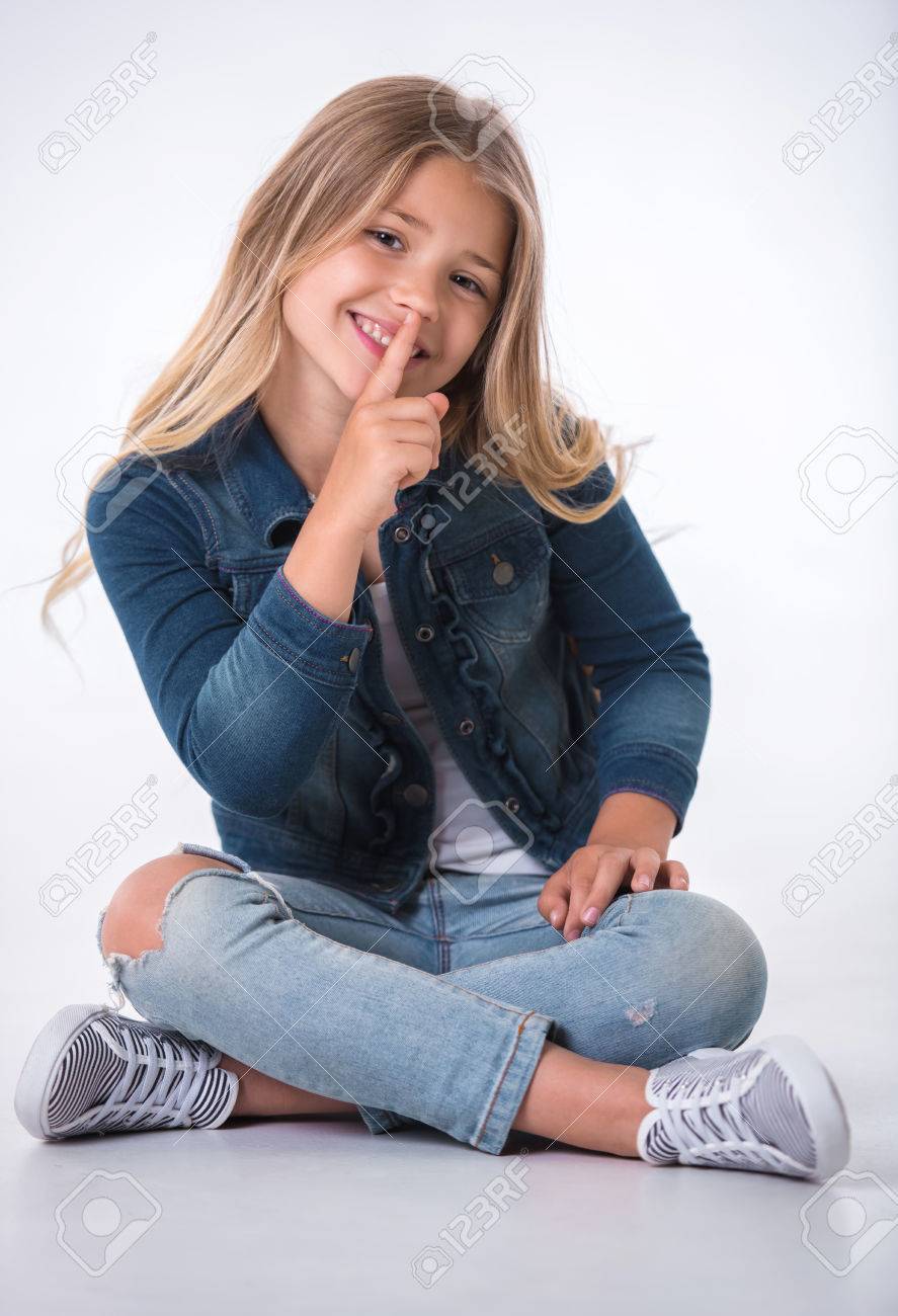 Beautiful little girl is showing silence sign, looking at camera and smiling while sitting on the floor on light background - 85605535 Beautiful little girl is showing silence sign, looking at camera and smiling while sitting on the floor on light background - 85605535