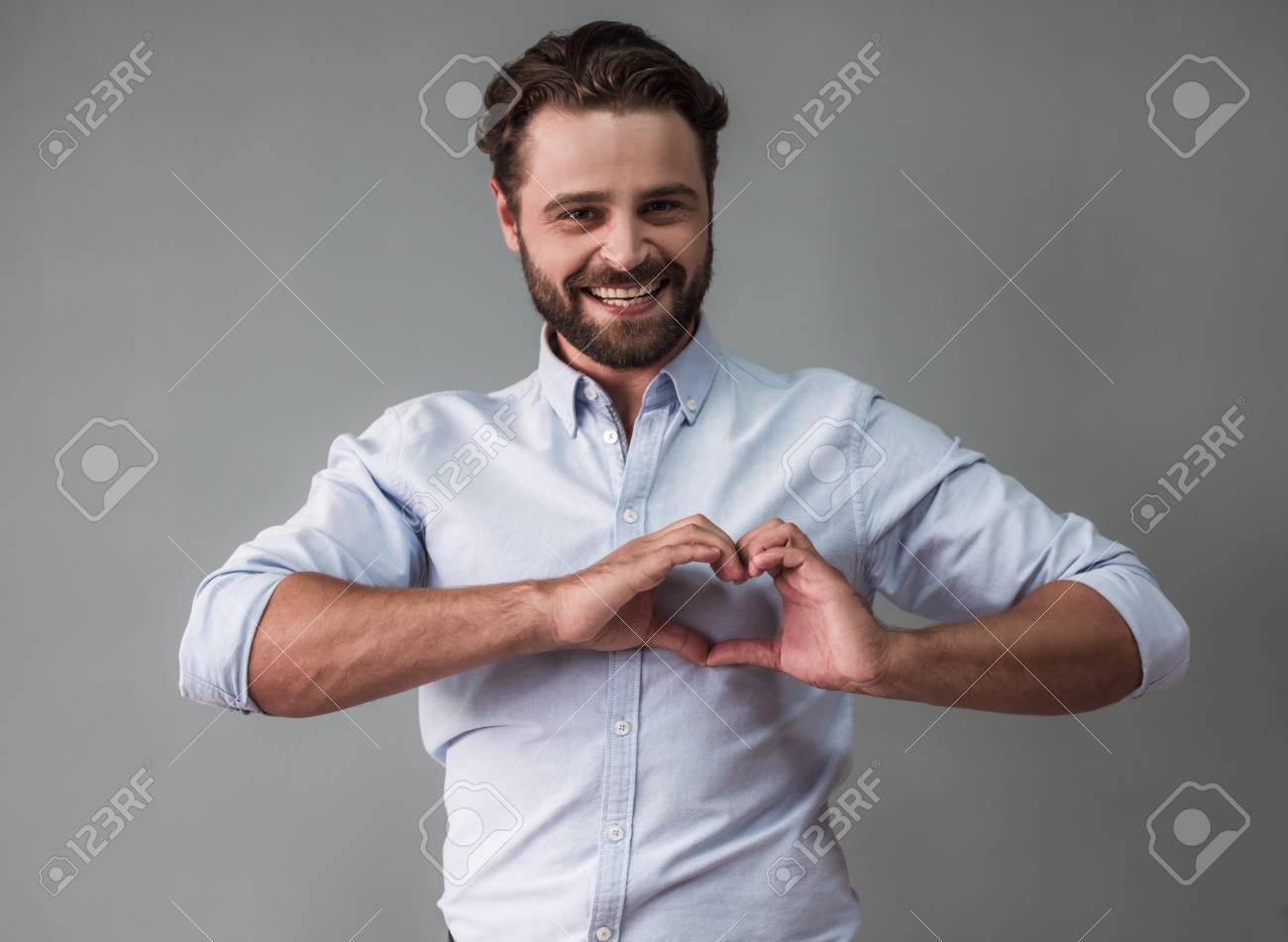 Handsome bearded businessman in smart casual wear is showing a heart, looking at camera and smiling, on gray background - 86472101 Handsome bearded businessman in smart casual wear is showing a heart, looking at camera and smiling, on gray background - 86472101