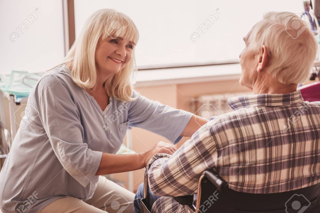 Beautiful old lady is talking to her husband and smiling while he is sitting in the wheelchair in hospital ward - 102322514 Beautiful old lady is talking to her husband and smiling while he is sitting in the wheelchair in hospital ward - 102322514
