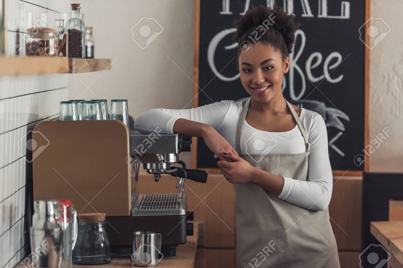 Beautiful Afro American barista in apron is looking at camera and smiling while leaning on the coffee machine - 107928401 Beautiful Afro American barista in apron is looking at camera and smiling while leaning on the coffee machine - 107928401