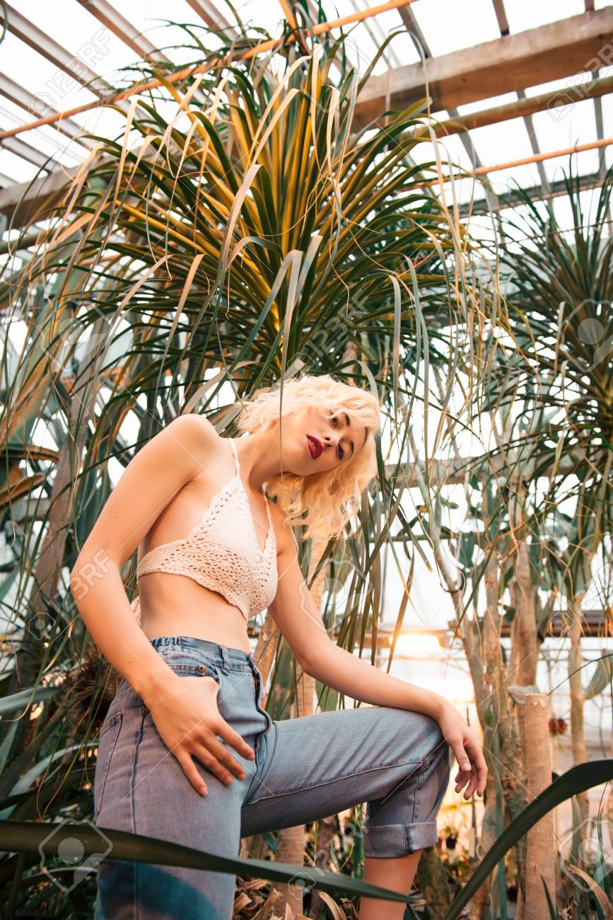 Young Perky Blonde Model With Perfect Body Shape, Clad In Sexy White Top  And Jeans, Teasing Camera With Lingering Glance. Posing Among Palm Trees In  A Greenhouse. Low Angle View. Stock Photo,