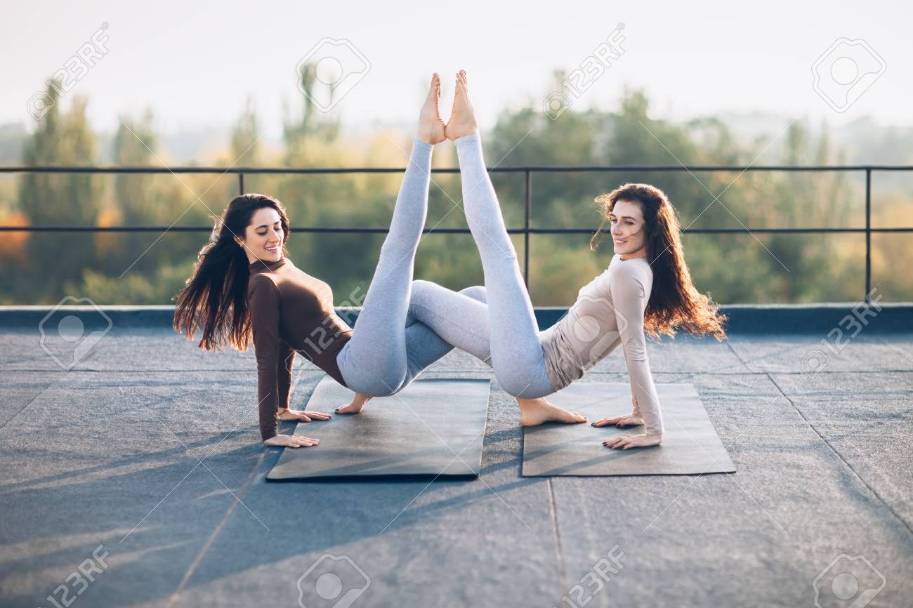 屋外の屋根の上でアクロヨガアダナをやっている2人の美しい女性 手を握った三角形の同期ポーズ 柔軟性 強さ 美しさ 集中力の概念 の写真素材 画像素材 Image