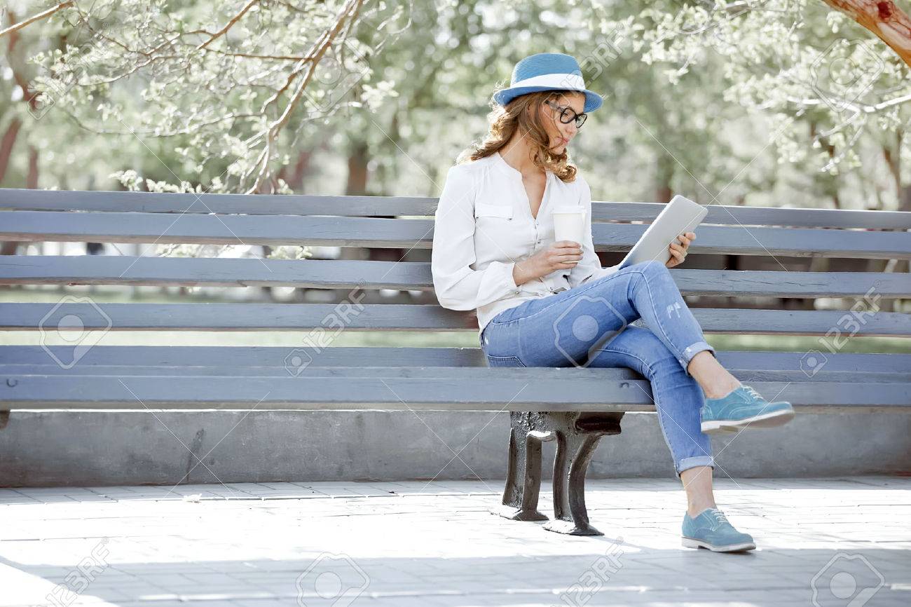 Happy young student with a tablet and a disposable coffee cup sitting on the bench and reading in a summer park. - 51083093 Happy young student with a tablet and a disposable coffee cup sitting on the bench and reading in a summer park. - 51083093