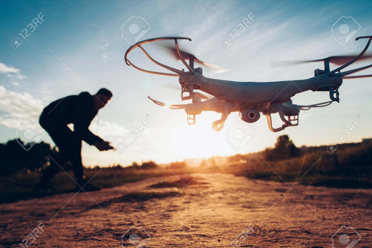 Drone trends and innovations. Modern hobby and leisure. Closeup of camera quadcopter controlled remotely by guy over blur sunset. - 121261053 Drone trends and innovations. Modern hobby and leisure. Closeup of camera quadcopter controlled remotely by guy over blur sunset. - 121261053