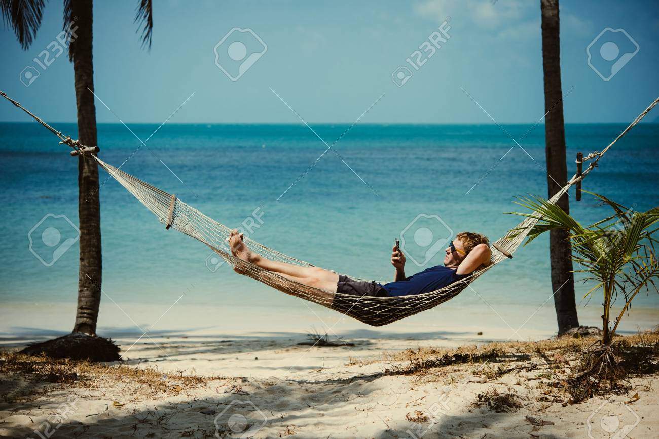 A young man relaxes in a hammock at the beach while checking messages on his smartphone. Toned image - 69677423 A young man relaxes in a hammock at the beach while checking messages on his smartphone. Toned image - 69677423