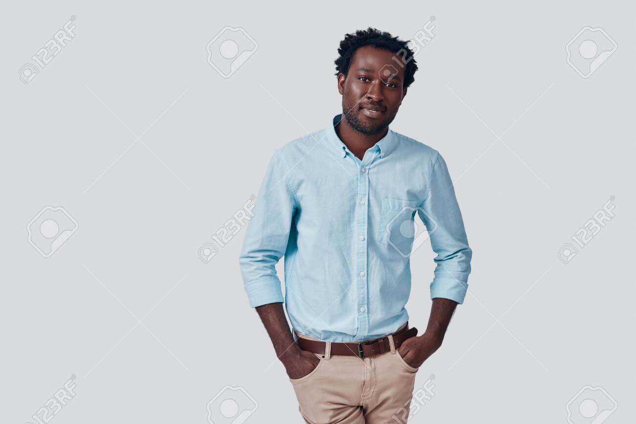 Handsome young African man looking at camera and smiling while standing against grey background - 135471605 Handsome young African man looking at camera and smiling while standing against grey background - 135471605