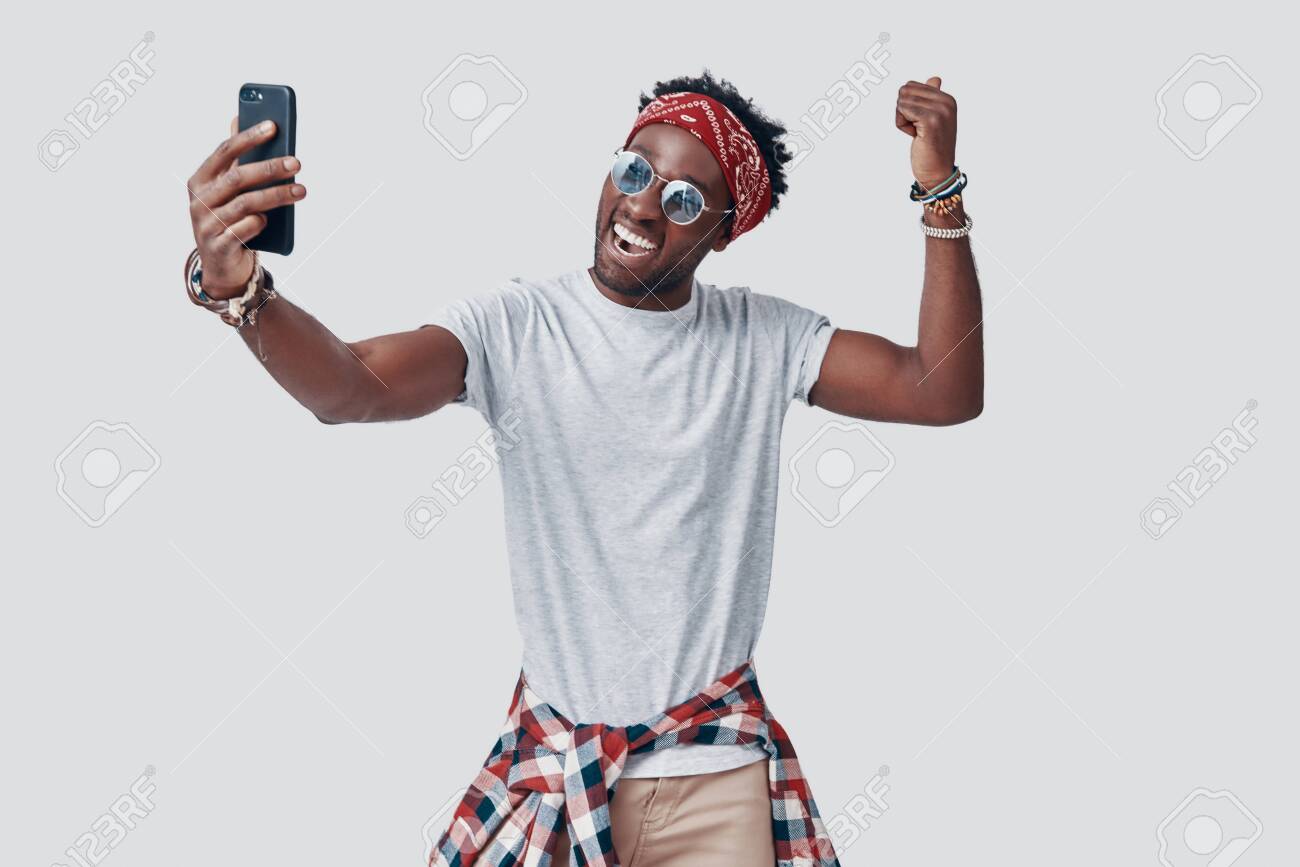Handsome young African man taking selfie and smiling while standing against grey background - 135471608 Handsome young African man taking selfie and smiling while standing against grey background - 135471608