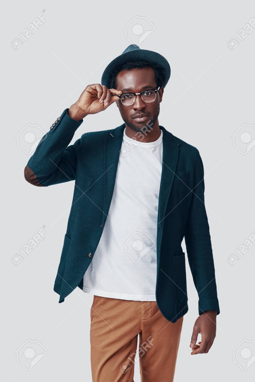Handsome young African man adjusting eyewear and looking at camera while standing against grey background - 135471625 Handsome young African man adjusting eyewear and looking at camera while standing against grey background - 135471625