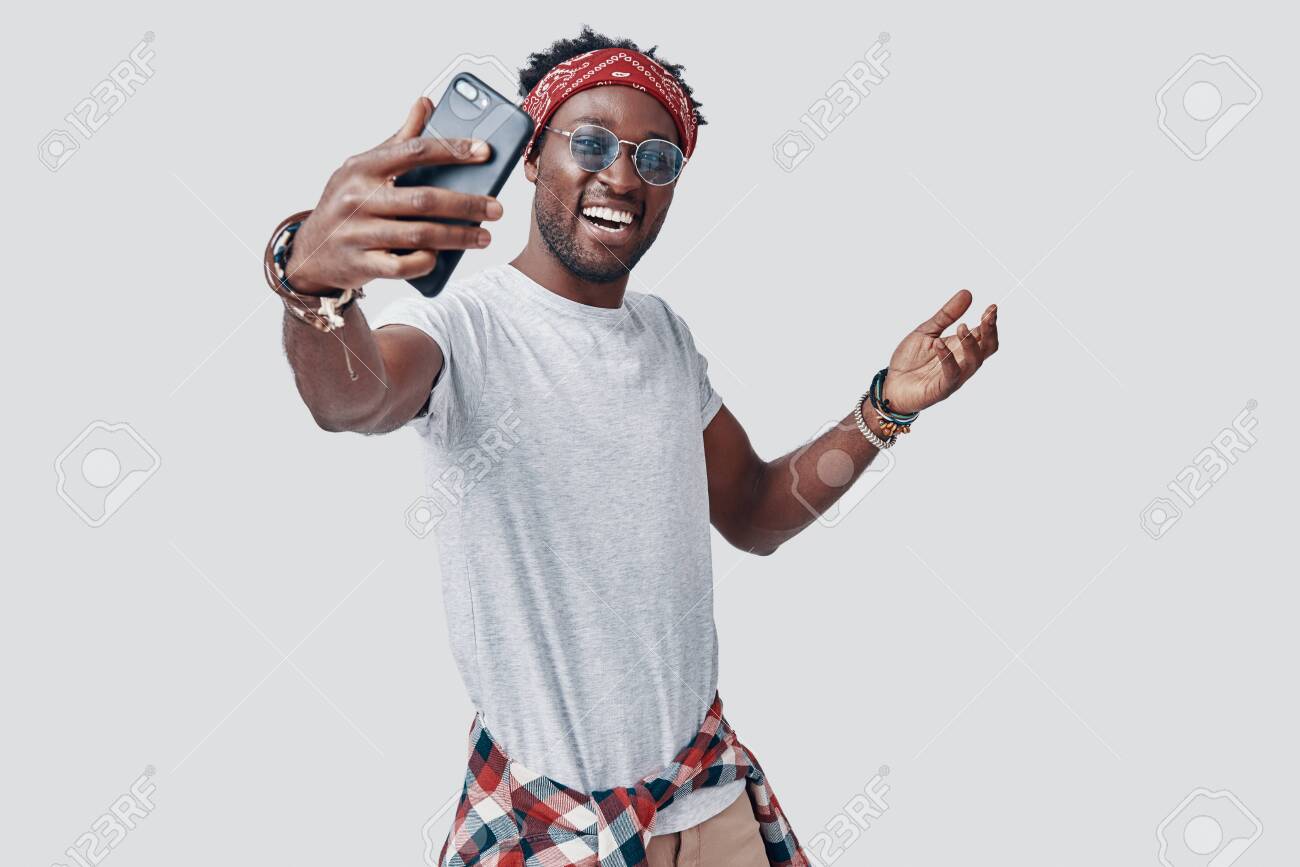 Handsome young African man taking selfie and smiling while standing against grey background - 135471642 Handsome young African man taking selfie and smiling while standing against grey background - 135471642