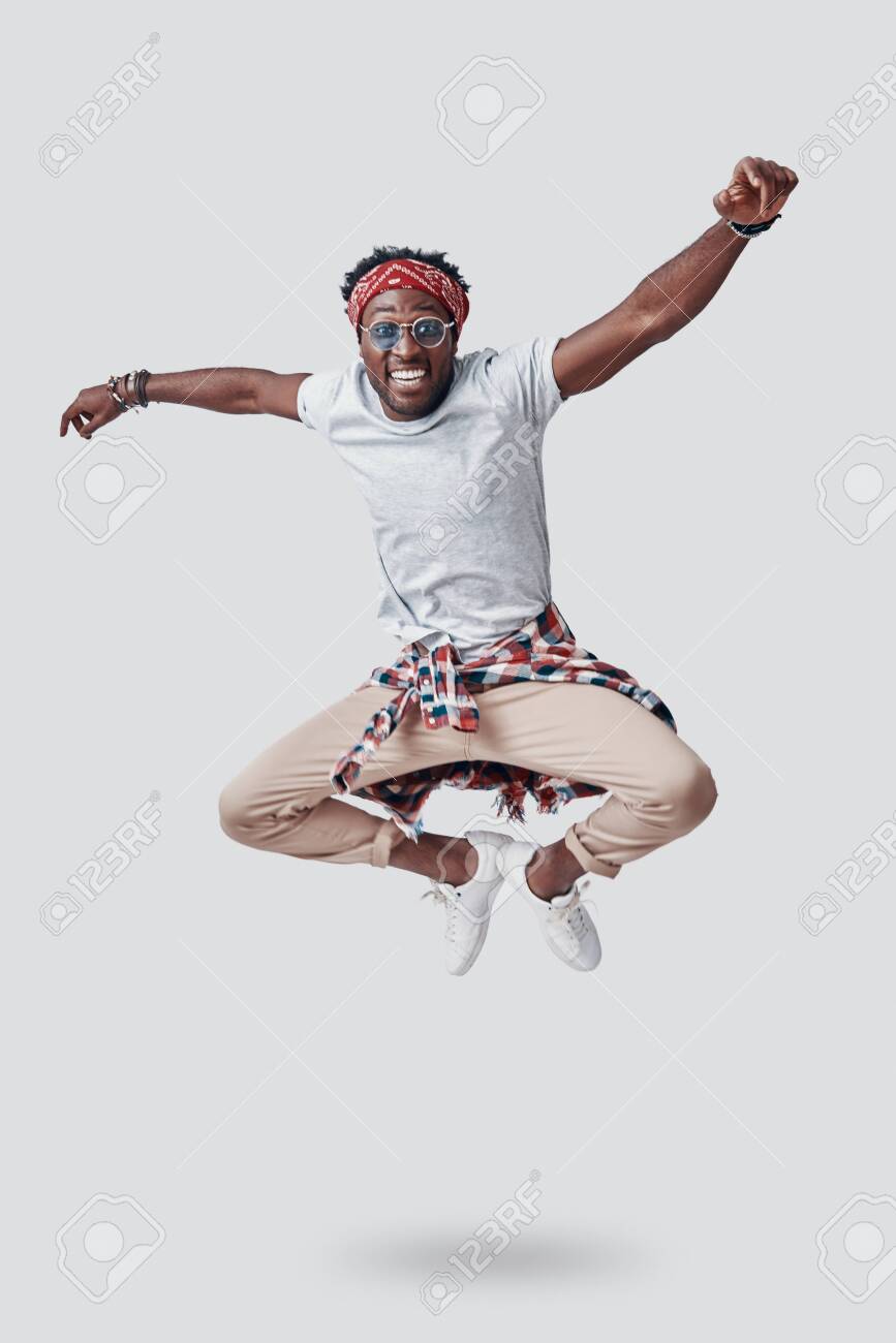 Full length of playful young African looking at camera and smiling while hovering against grey background - 135471644 Full length of playful young African looking at camera and smiling while hovering against grey background - 135471644