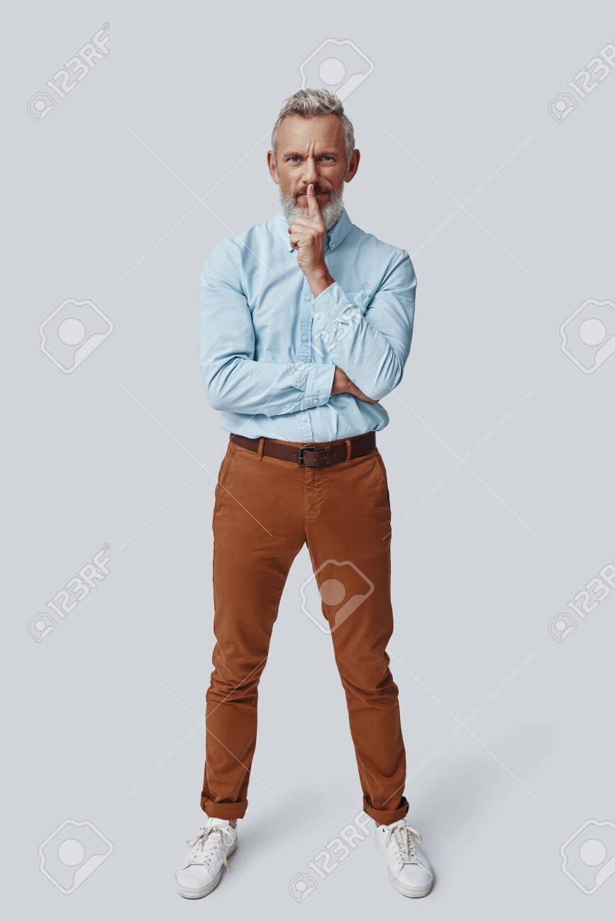 Full length of mature man looking at camera and keeping finger on lips while standing against grey background - 138444909 Full length of mature man looking at camera and keeping finger on lips while standing against grey background - 138444909