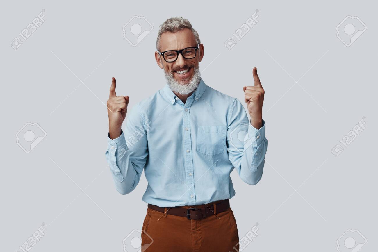Cheerful mature man smiling and pointing copy space while standing against grey background - 138444943 Cheerful mature man smiling and pointing copy space while standing against grey background - 138444943