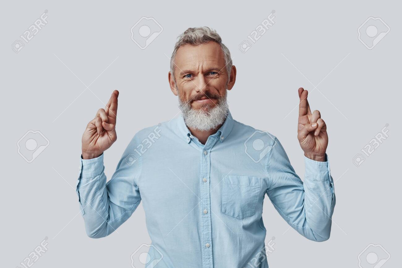 Mature man looking at camera and keeping fingers crossed while standing against grey background - 137917217 Mature man looking at camera and keeping fingers crossed while standing against grey background - 137917217